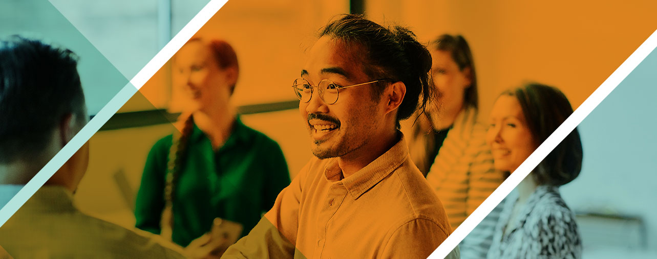 A man wearing glasses is smiling, while sitting among a group of coworkers.