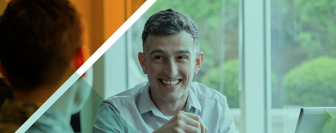 A young, white man is smiling, while having a conversation with someone in a business setting.