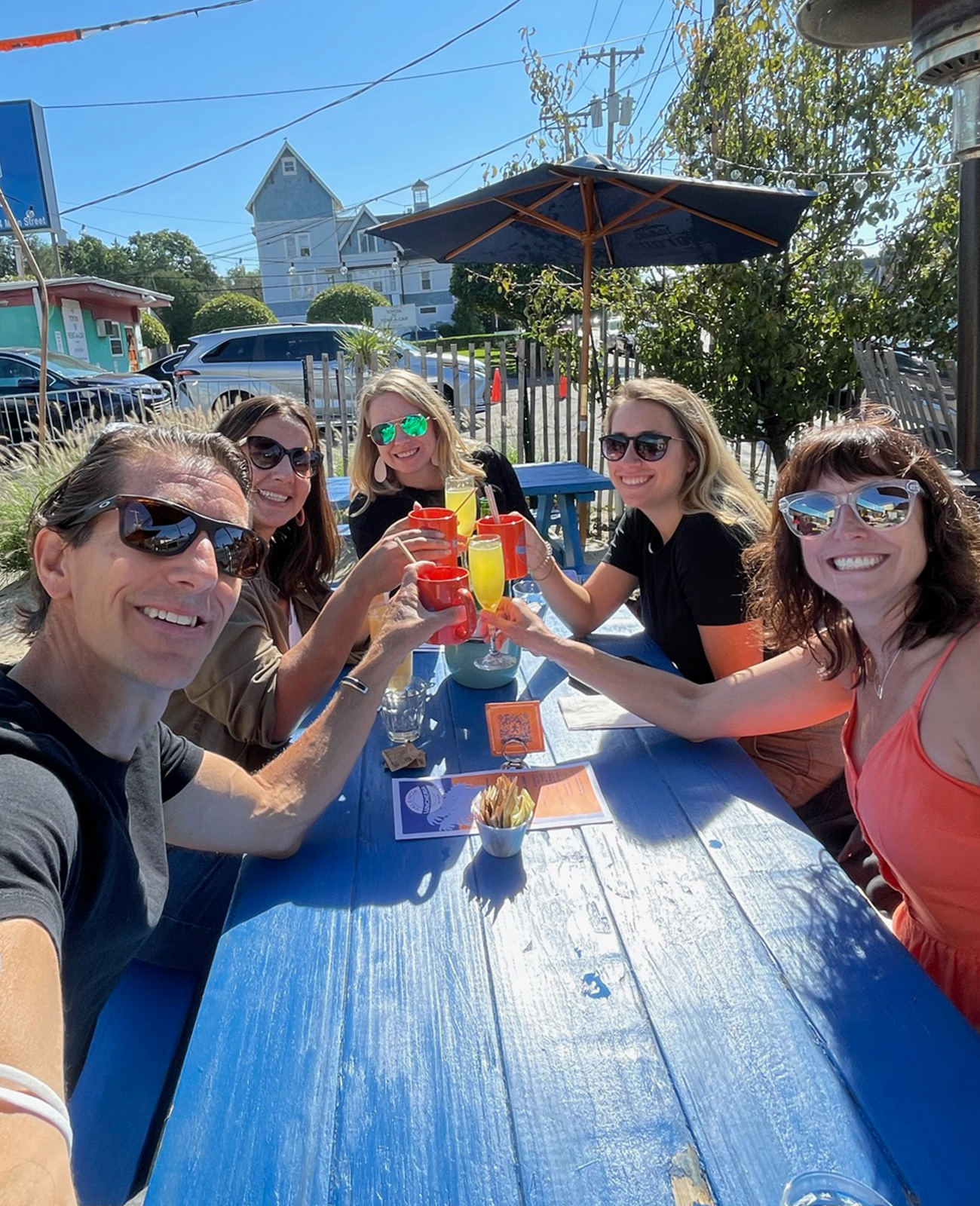 Five Ingenuity Design colleagues smiling and raising their glasses together at an outdoor table. 