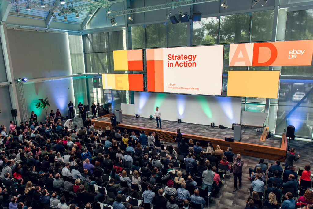 A presenter speaking in a massive auditorium below large screens displaying custom strategy and action graphics for eBay. 
