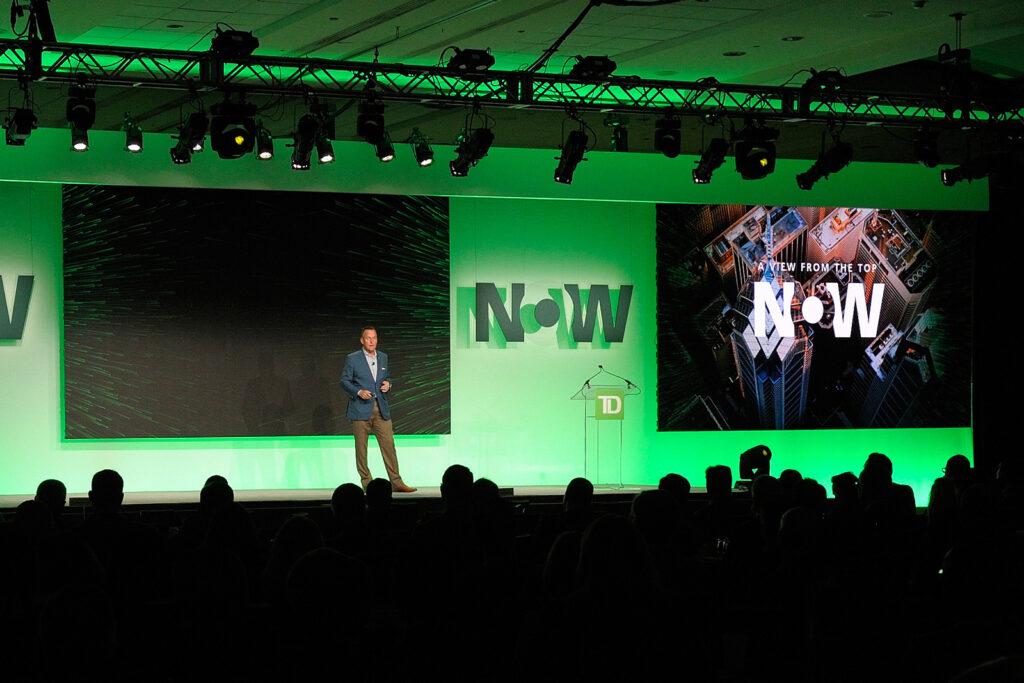 A keynote presenter speaking on a TD Bank stage beneath a large green neon sign in front of an audience.