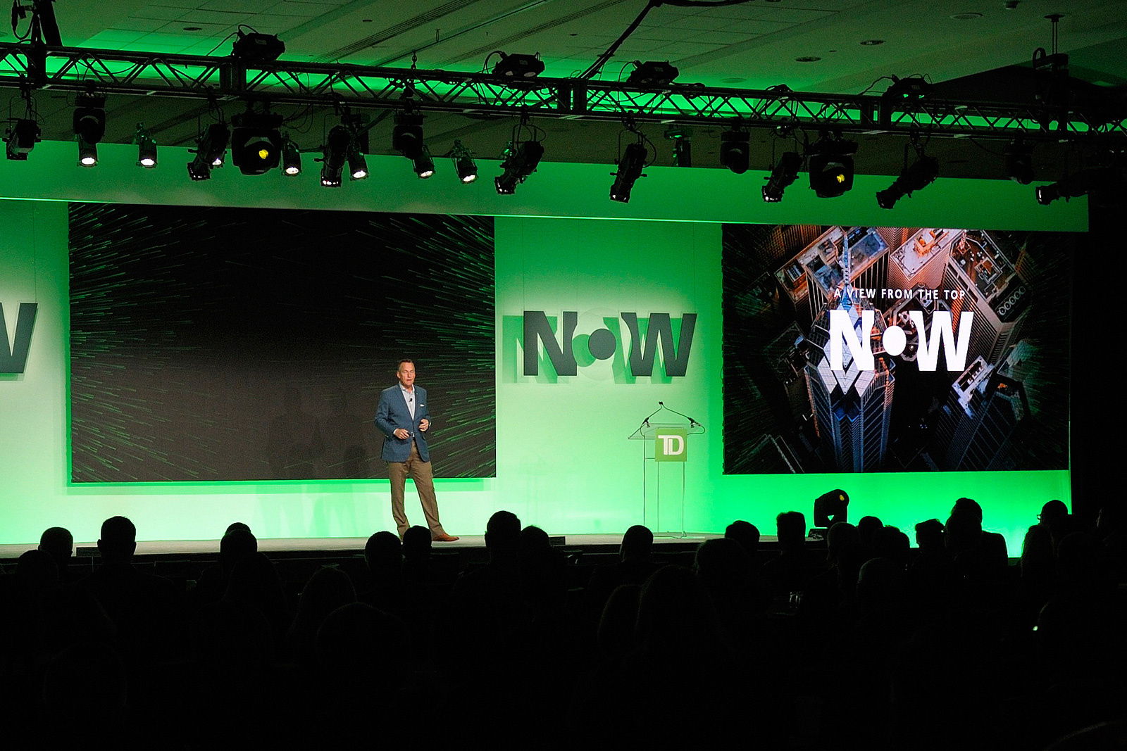 A keynote presenter speaking on a TD Bank stage beneath a large green neon sign in front of an audience.