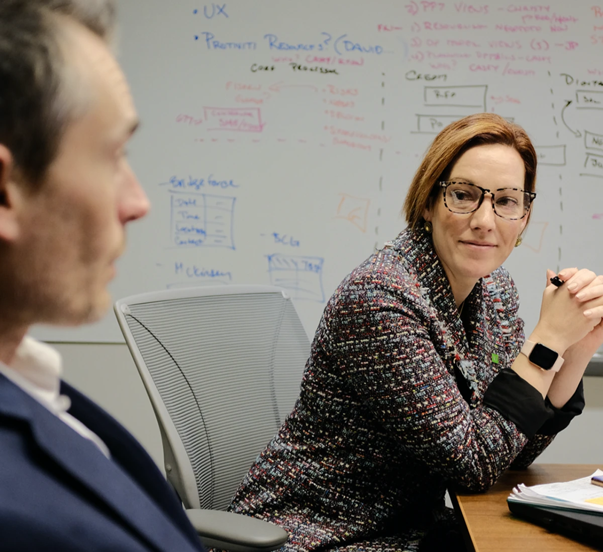 A businesswoman wearing glasses in a meeting, sitting in front of a whiteboard filled with strategy notes.