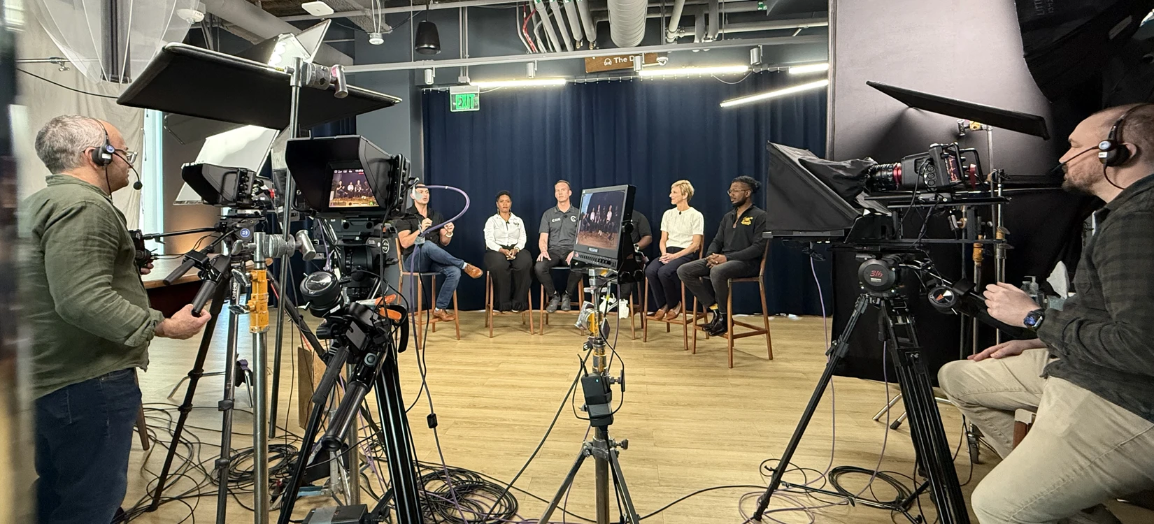 A broad view of a video production set with cameras and lighting rigs filming a multi-speaker panel.
